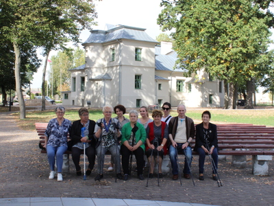 Zdjęcie ukazuje seniorów i opiekunów siedzących na ławce w parku. W tle pałac we Wierzbinku.