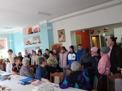 Zdjęcie wykonane w sali głównej Domu Dziennego Pobytu dla Osób Starszych w Sompolnie. Przedstawia grupę dzieci  wraz z seniorami.