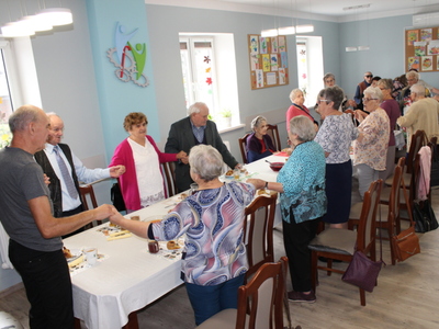 Zdjęcie wykonane w sali głównej Domu Dziennego Pobytu dla osób starszych w Sompolnie. Przedstawia seniorów stojących przy stołach, trzymających się za ręce i śpiewających piosenkę.