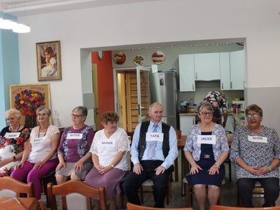 Na zdjęciu seniorzy siedzący w rzędzie na krzesłach, przygotowują się do zabawy w Rodzinę. W tle pracownia kulinarna Domu Dziennego Pobytu.