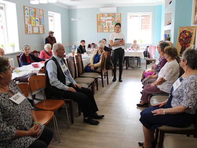 Na fotografii seniorzy podczas zabawy w rodzinę. W tle osoby starsze siedzące przy stołach.