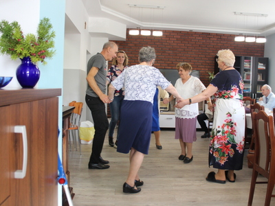 Zdjęcie przedstawia seniorów wraz z opiekunami tańczących na sali głównej DDP, z lewej strony zdjęcia niebieski wazon z gałązkami cisu.