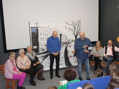 1)	Zdjęcie przedstawia wnętrze kina za rogiem w Sompolnie. Na forum widać seniorów – aktorów siedzących na ławeczkach oraz dwóch mężczyzn przemawiających do publiczności.