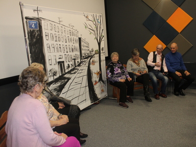 3)	Zdjęcie przedstawia seniorów grających swoje role. Po środku scenografia przedstawiająca miejskie bloki.