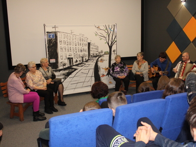 4)	Zdjęcie przedstawia seniorów grających swoje role. Po środku scenografia przedstawiająca miejskie bloki. Jeden mężczyzna gra na gitarze a drugi na akordeonie.