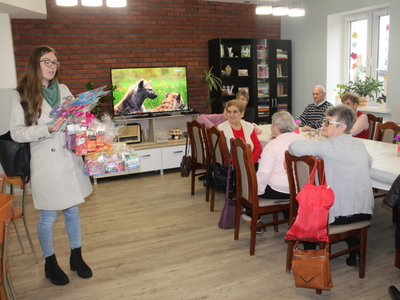 Zdjęcie przedstawia nauczycielkę przedszkola, która czyta życzenia oraz przekazuje kosze z podarunkami  dla seniorów.