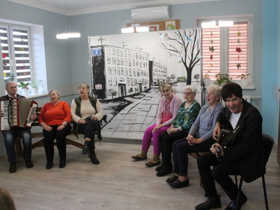 Zdjęcie przedstawia seniorów grających swoje role. Po środku scenografia przedstawiająca miejskie bloki. Jeden mężczyzna gra na gitarze a drugi na akordeonie.