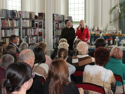 2)	Fotografia przedstawia dwie kobiety witające osoby zebrane na otwarciu wystawy. W tle rękodzieło oraz półki z książkami.