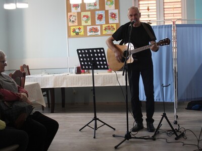 1)	Zdjęcie przedstawia Pana Andrzeja Prętnickiego stojącego przy mikrfonie na sali głównej DDP. W rękach trzyma gitarę.