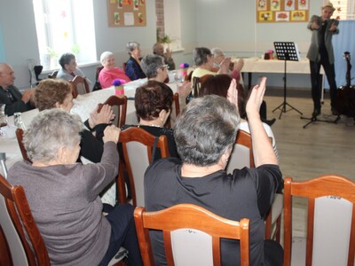 2)	Na zdjęciu widać publiczność składającą się z Seniorów z DDP, którzy biją brawa. Na drugim planie występujący przed nimi – Pan Andrzej Prętnicki