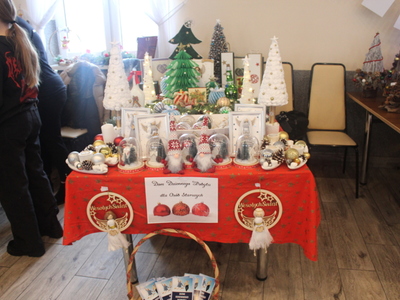 Zdjęcie wykonane w sali OSP Sompolno. Ukazuje stoisko Domu Dziennego Pobytu dla osób starszych w Sompolnie. Na czerwonym obrusie stoją ozdoby świąteczne