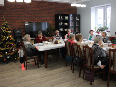 2)	Na zdjęciu seniorzy siedzący przy stole. Po lewej stronie widoczna przystrojona choinka.
