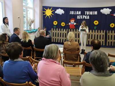 1)	Zdjęcie wykonane w jednej z sal szkoły w Mąkolnie. Z tyłu granatowa gazetka rozmieszczona na całej ścianie. Na niej płot, strach na wróble, słoneczniki i napis Julian Tuwim „Rzepka”. Na tle gazetki dziewczyna z mikrofonem. Z przodu widownia siedząca na krzesłach.