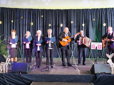 Fotografia wykonana w hali sportowej zespołu szkolno – przedszkolnego w Lubstowie podczas samorządowego kolędowania w gminie Sompolno. Przedstawia scenę na której występują seniorzy z domu dziennego pobytu w Sompolnie, Pan Tomasz Majewski - dyrektor miejsko – gminnego ośrodka pomocy społecznej w Sompolnie oraz Pan Andrzej Prętnicki – instruktor muzykoterapii oraz radny rady miejskiej w Sompolnie.