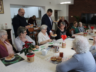 Zdjęcie wykonane na sali głównej domu dziennego pobytu w Sompolnie. Widać na nim burmistrza miasta Sompolna i dyrektora mgops w Sompolnie roznoszących pączki seniorom siedzącym przy stole