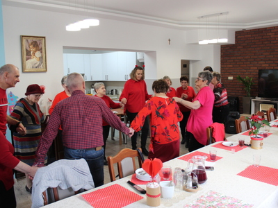 Zdjęcie przedstawia seniorów wraz z opiekunami tańczących w kółku. Wszyscy posiadają na sobie chociaż jeden element stroju w kolorze czerwonym.