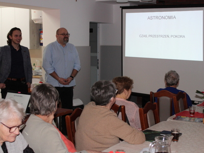 Zdjęcie wykonane na sali głównej Domu Dziennego Pobytu dla Osób Starszych w Sompolnie. Widać na nim dyrektora MGOPS w Sompolnie oraz Pana Mikołaja Błaszczyka – ucznia liceum w Sompolnie. Po prawej stronie tytuł prezentacji – ASTRONOMIA CZAS PRZESTRZEŃ POKORA. Seniorzy siedzący na krzesłach przy stole.