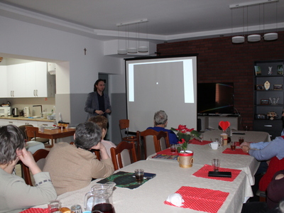 Na zdjęciu seniorzy siedzący przy stole. W tle Pan Mikołaj Błaszczyk prezentujący zdjęcie własnego autorstwa.