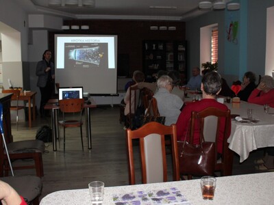 Na fotografii seniorzy siedzący przy stole w sali głównej DDP. W tle ekran z tytułem slajdu KRÓTKA HISTORIA. Po prawej stronie seniorzy siedzący przy stole.