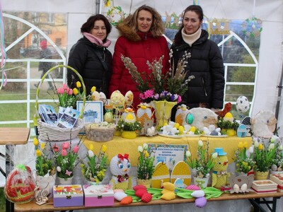 1)	Zdjęcie przedstawia stoisko Domu Dziennego Pobytu podczas Sompoleńskiego Kiermaszu Wielkanocnego. Przy stoisku stoją trzy pracownice Ośrodka.