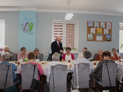 2)	Na fotografii jeden z seniorów stojąc za stołem, przemawia podczas spotkania wielkanocnego. Przy stole siedzą wszyscy uczestnicy DDP.
