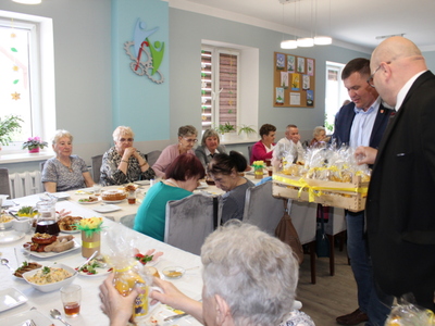 3)	Na zdjęciu dyrektor wraz z burmistrzem wręczają seniorom siedzącym za stołem świąteczne upominki.