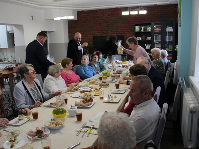 4)	Zdjęcie wykonane w sali głównej domu dziennego pobytu, przedstawia dyrektora wraz z burmistrzem wręczających seniorom siedzącym za stołem świąteczne upominki.