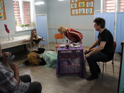 1)	Zdjęcie wykonane w sali głównej domu dziennego pobytu w Sompolnie. Przedstawia scenę z komedii pt. „Czekoladki”. Aktor w stroju kobiety leży na ziemi, a młoda kobieta przegląda jego torebkę. Inna aktorka pochyla się nad leżącym, przy stole siedzi czwarty aktor.