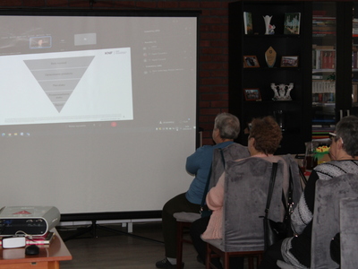 2)	Na zdjęciu seniorzy oglądający prezentację o cyberbezpieczeństwie. W tle ekran z wyświetlanym slajdem, z lewej strony rzutnik.