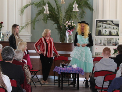 2)	Na głównym planie zdjęcia aktorzy grający w sztuce: dwie kobiety i mężczyzna przebrany za kobietę. Po obu stronach sceny publiczność oglądająca przedstawienie.