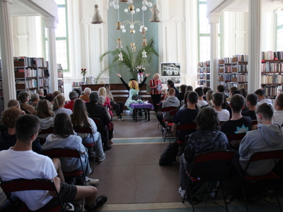 3)	Na zdjęciu aktorzy grający w sztuce pt. „Czekoladki”. Mężczyzna przebrany za kobietę wznosi ręce do góry. Po obu stronach sceny publiczność oglądająca przedstawienie oraz widoczne regały z książkami.