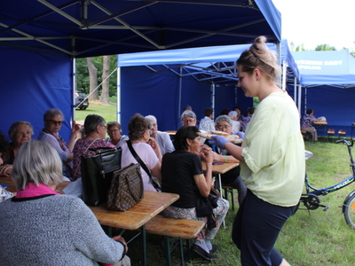 1.  Zdjęcie przedstawia grupę Seniorów siedzących pod namiotami i Pracownice MGOPS serwującą chleb ze smalcem.