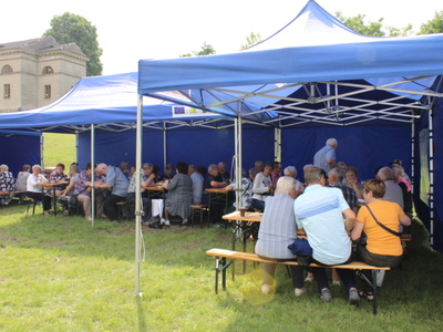 7. Fotografia przedstawia seniorów siedzących pod niebieskimi namiotami podczas majówki seniorów.