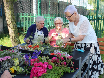 1)	Zdjęcie wykonane na terenie zielonym przy Miejsko – Gminnym Ośrodku Pomocy Społecznej w Sompolnie. Przedstawia trzy seniorki które plotą wianek.