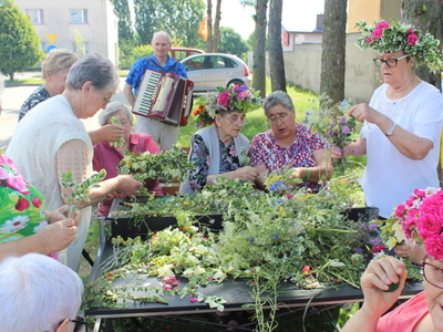 2)	Na zdjęciu grupa seniorek plecie wianki. W tle uczestnik DDP z akordeonem.