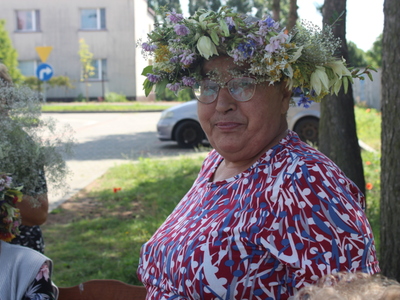 5)	Na zdjęciu seniorka w kolorowej bluzce i wianku pozująca do zdjęcia.