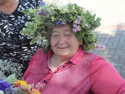 6)	Fotografia przedstawia starszą kobietę w różowej bluzce i wianku pozującą do zdjęcia.