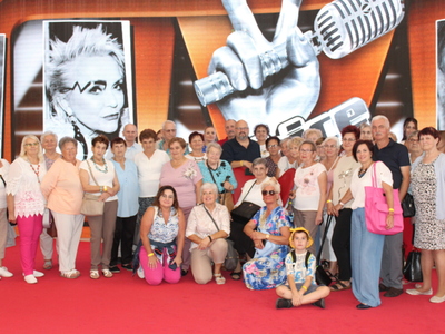 4)	Zdjęcie grupowe wykonane w siedzibie telewizji polskiej w Warszawie. Przedstawia grupę seniorów z  Sompolna i okolic wraz z opiekunami pozującą na tle plakatu THE VOICE SENIOR.