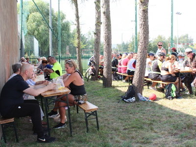 5)	Na zdjęciu uczestnicy rajdu wraz z seniorami wspólnie siedzący przy stołach na świeżym powietrzu i spożywający posiłek.