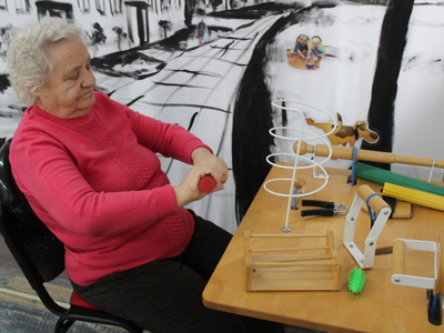 3.	Zdjęcie przedstawia starszą kobietę wykonującą ćwiczenia rąk przy stoliku do terapii manualnej ręki.