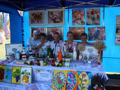 1)	Zdjęcie wykonane pod namiotem domu dziennego pobytu podczas dożynek powiatowych w Lubstowie. Przedstawia grupę uczestników DDP. W tle obrazy, a z przodu wystawa rękodzieła.