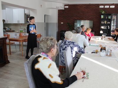 Zdjęcie wykonane w sali głównej Domu Dziennego Pobytu Senior+ w Sompolnie. Przedstawia Panią Joannę Grzechowiak – doradcę obywatelskiego mówiącą do seniorów siedzących przy stole.