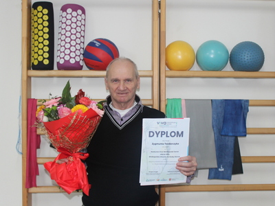 5)	Zdjęcie wykonane w sali rehabilitacyjnej przedstawia seniora trzymającego kwiaty i dyplom.