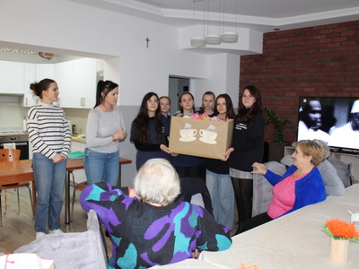 1)	Zdjęcie wykonane w sali głównej Domu Dziennego Pobytu Senior+ w Sompolnie. Przedstawia wolontariuszki ze szkoły w Mąkolnie trzymające karton z „Herbatką dla Seniora” oraz dwie panie nauczycielki. Przy stole siedzą seniorzy.