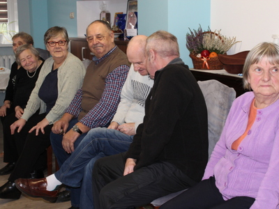 Na zdjęciu siedmioro seniorów siedzących na krzesłach i  uczestniczących w zabawie „głuchy telefon”.