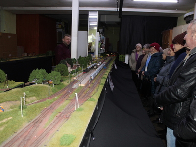 Zdjęcie wykonane przy makiecie kolei. Przedstawia seniorów oraz pana opowiadającego o transporcie kolejowym – właściciela makiety.