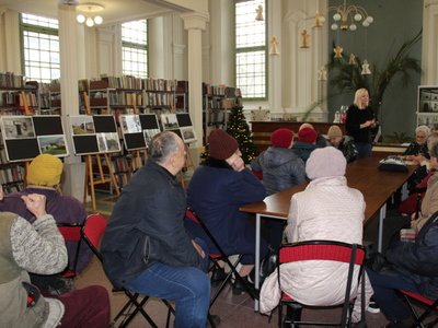 Zdjęcie wykonane w bibliotece. Przedstawia dyrektorkę biblioteki i seniorów siedzących przy stole. Z lewej strony wystawa fotografii  sompoleńskiej kolei wąskotorowej,