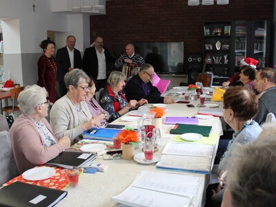 1)	Zdjęcie wykonane na sali głównej domu dziennego pobytu. Przedstawia seniorów siedzących przy stole, panią sekretarz, pana burmistrza, dyrektora mgops oraz jednego z uczestników stojącego z akordeonem.