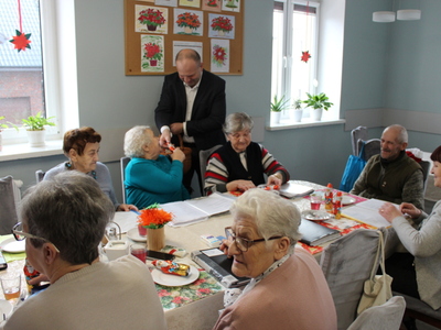 3)	Fotografia przedstawia burmistrza rozdającego czekoladowe mikołaje seniorom siedzącym przy stole.