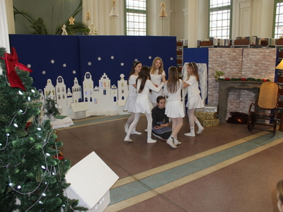 2)	Na zdjęciu dziewczynka siedząca na podłodze wokół której tańczą dziewczynki ubrane w białe stroje.